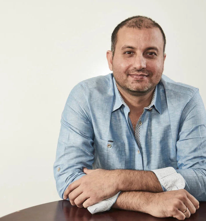 Kapil wearing a light blue shirt with rolled-up sleeves, sitting at a table against a plain background